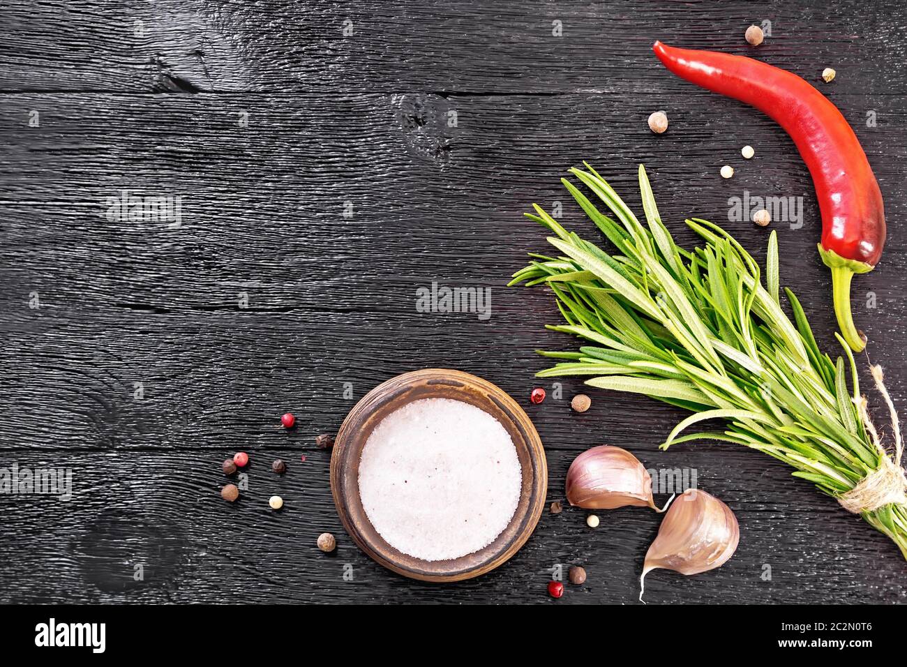 Telaio della rosa Sale Himalayano nel recipiente, i piselli di pepe colorati, un mazzetto di rosmarino fresco, un pod di peperoncino e due spicchi di aglio sul nero woo Foto Stock