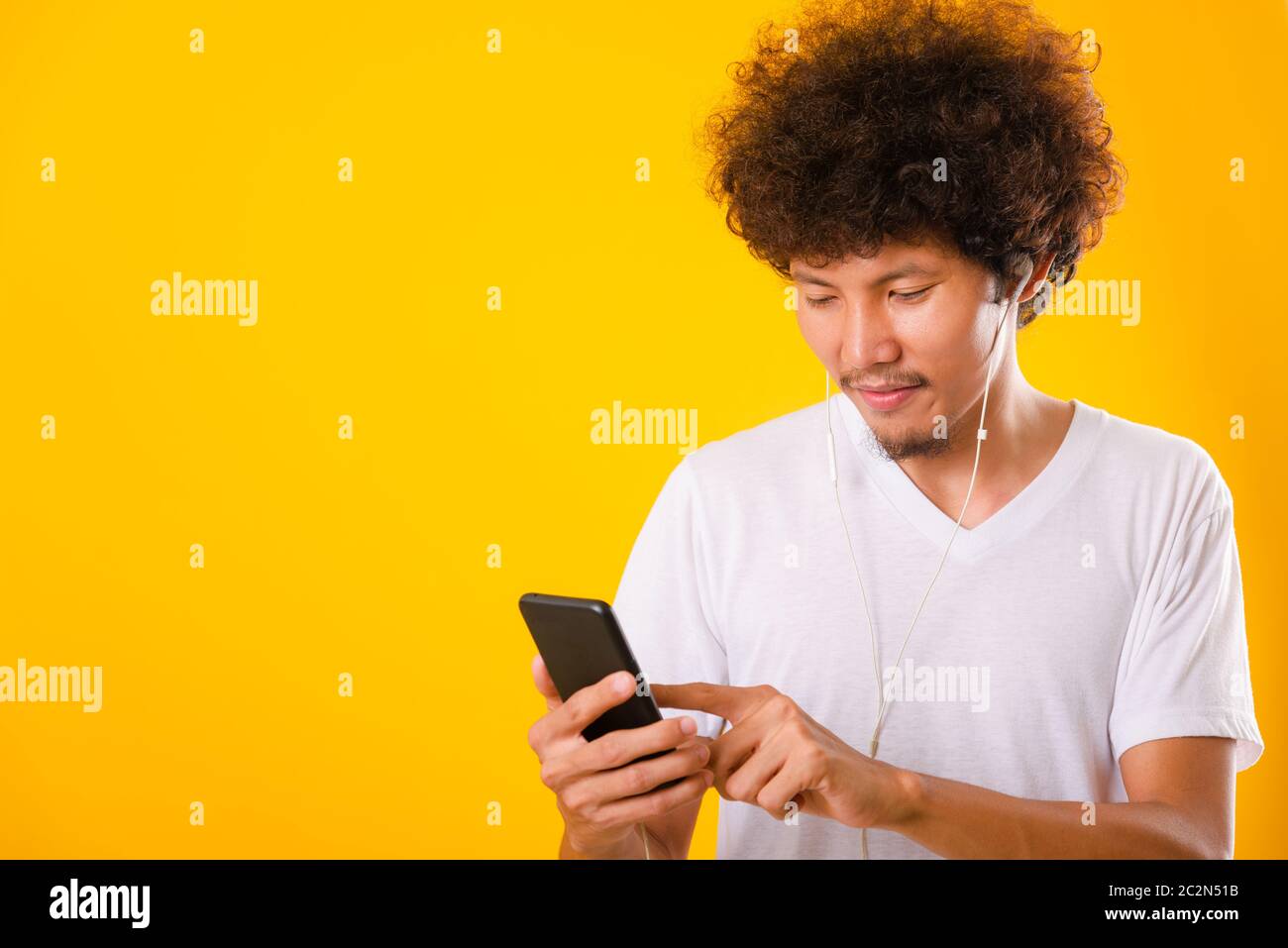Felice asiatico bello uomo con capelli ricci sorridente godendo di ascoltare la musica sulle cuffie utilizzando uno smartphone cellulare isolato su sfondo giallo Foto Stock