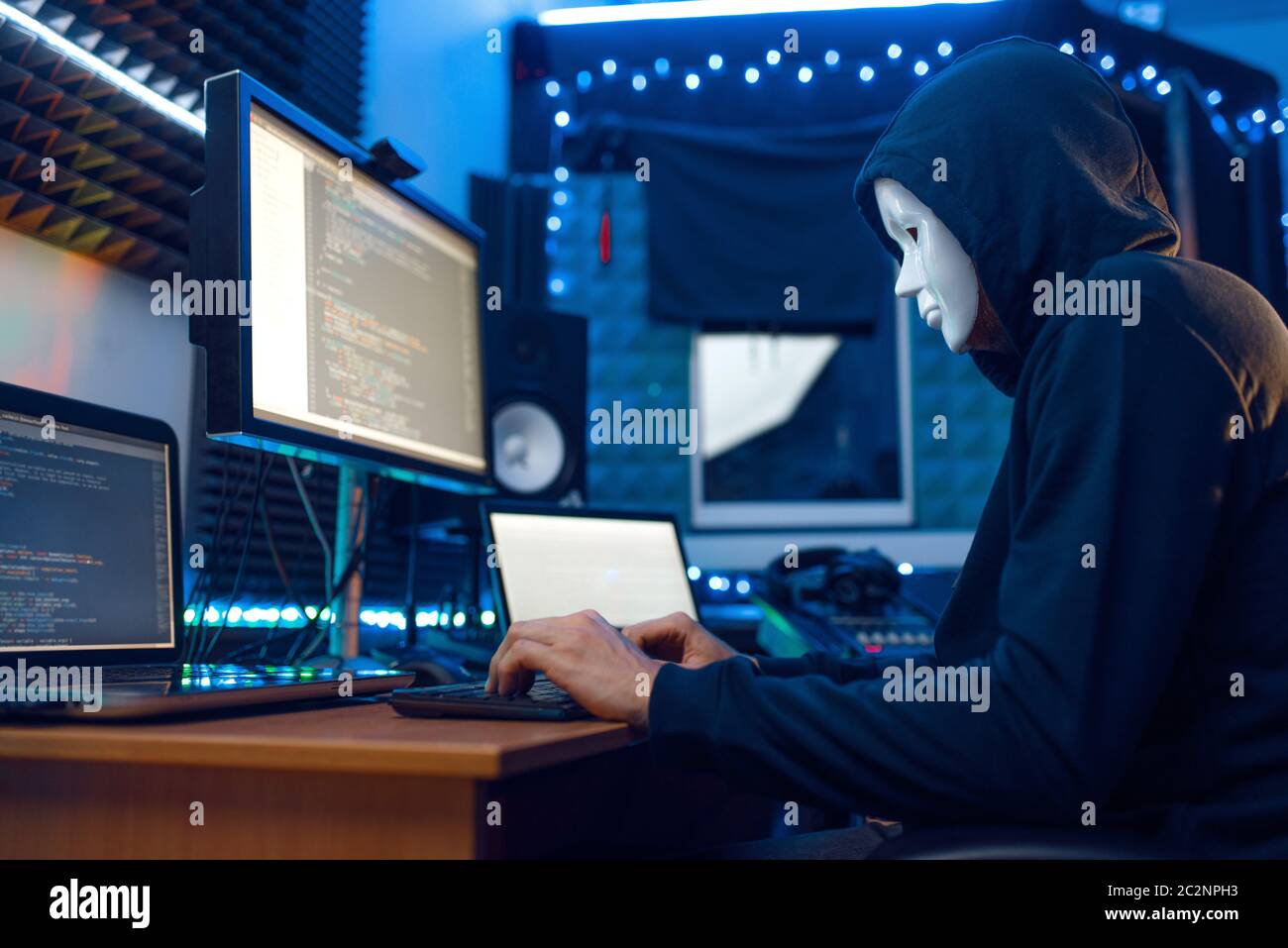 Hacker in maschera e cappuccio seduto sul suo posto di lavoro con laptop e PC, password o account hacking. Internet spia, stile di vita illegale, lavoro di rischio, rete c Foto Stock