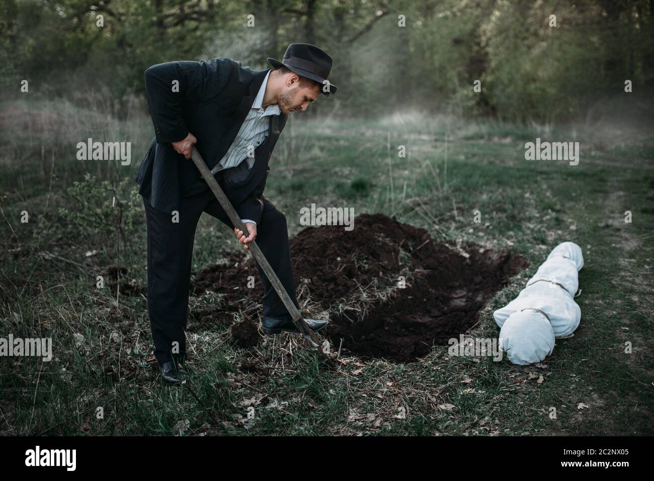 L'assassino maschile con una pala sta scavando una tomba per la vittima, il corpo avvolto in una tela, concetto maniaco seriale Foto Stock