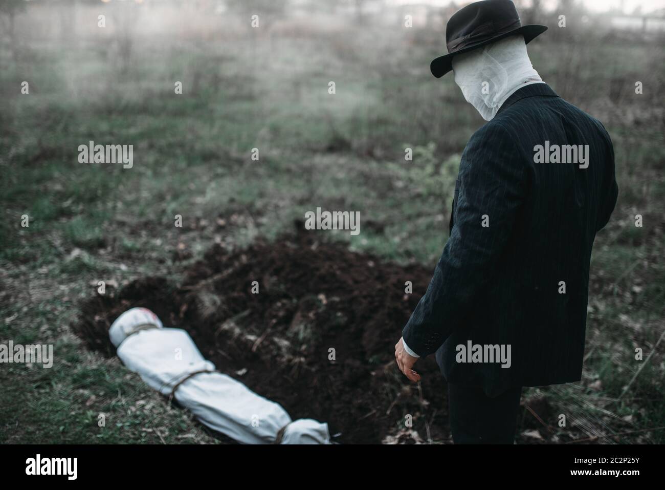 Bloody assassino si erge contro una tomba con la vittima nella foresta, il corpo avvolto in una tela, serial maniac concept Foto Stock