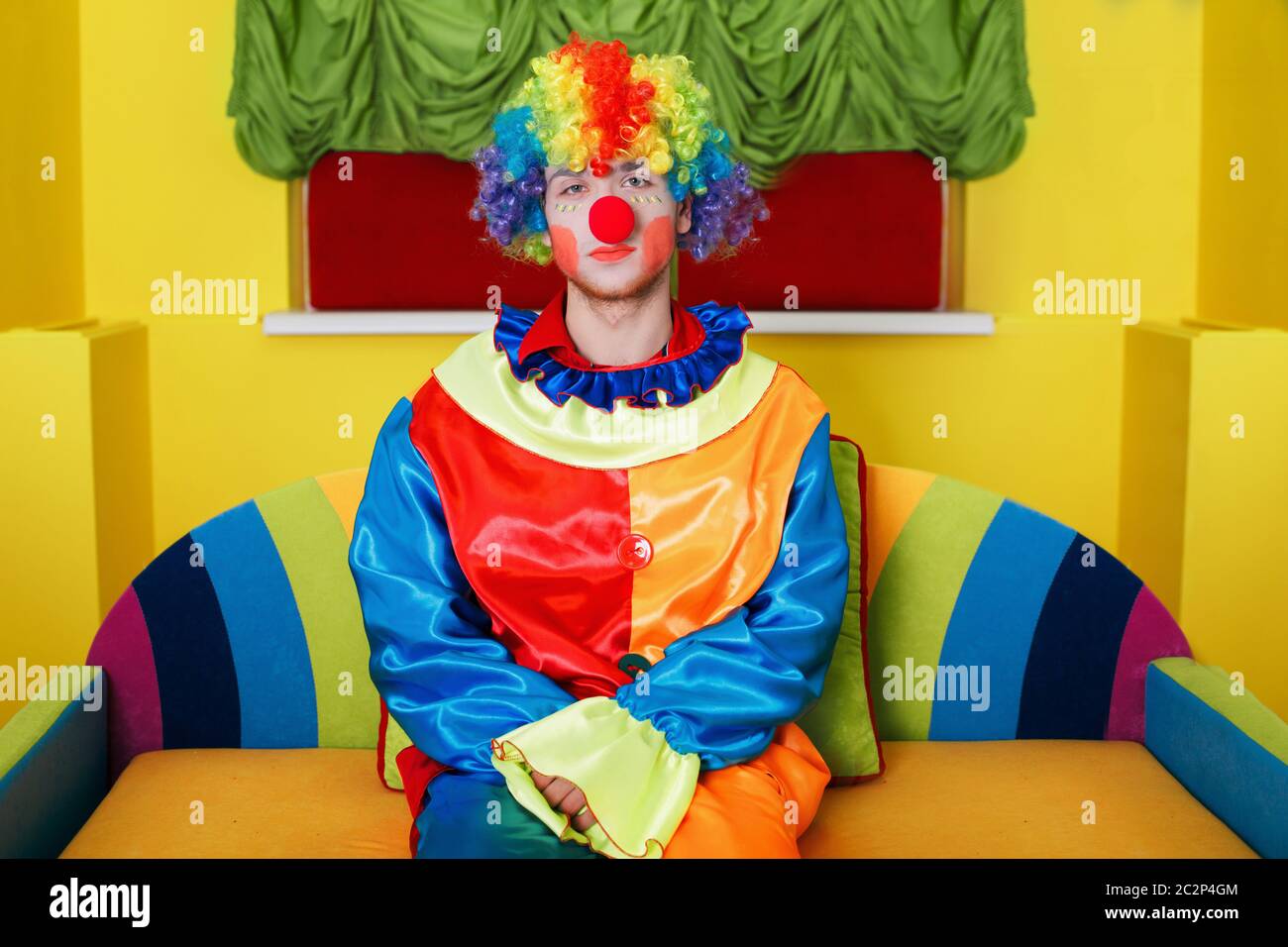 Clown in costume arcobaleno colorato con trucco e naso rosso seduto su divano colorato. Foto Stock