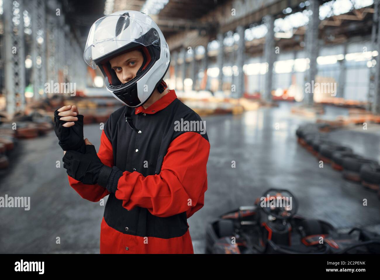 Kart racer in uniforme rossa, casco e guanti, kart auto sport indoor. Gara di velocità su pista da go-kart chiusa con barriera per pneumatici. Concorrenza rapida dei veicoli Foto Stock