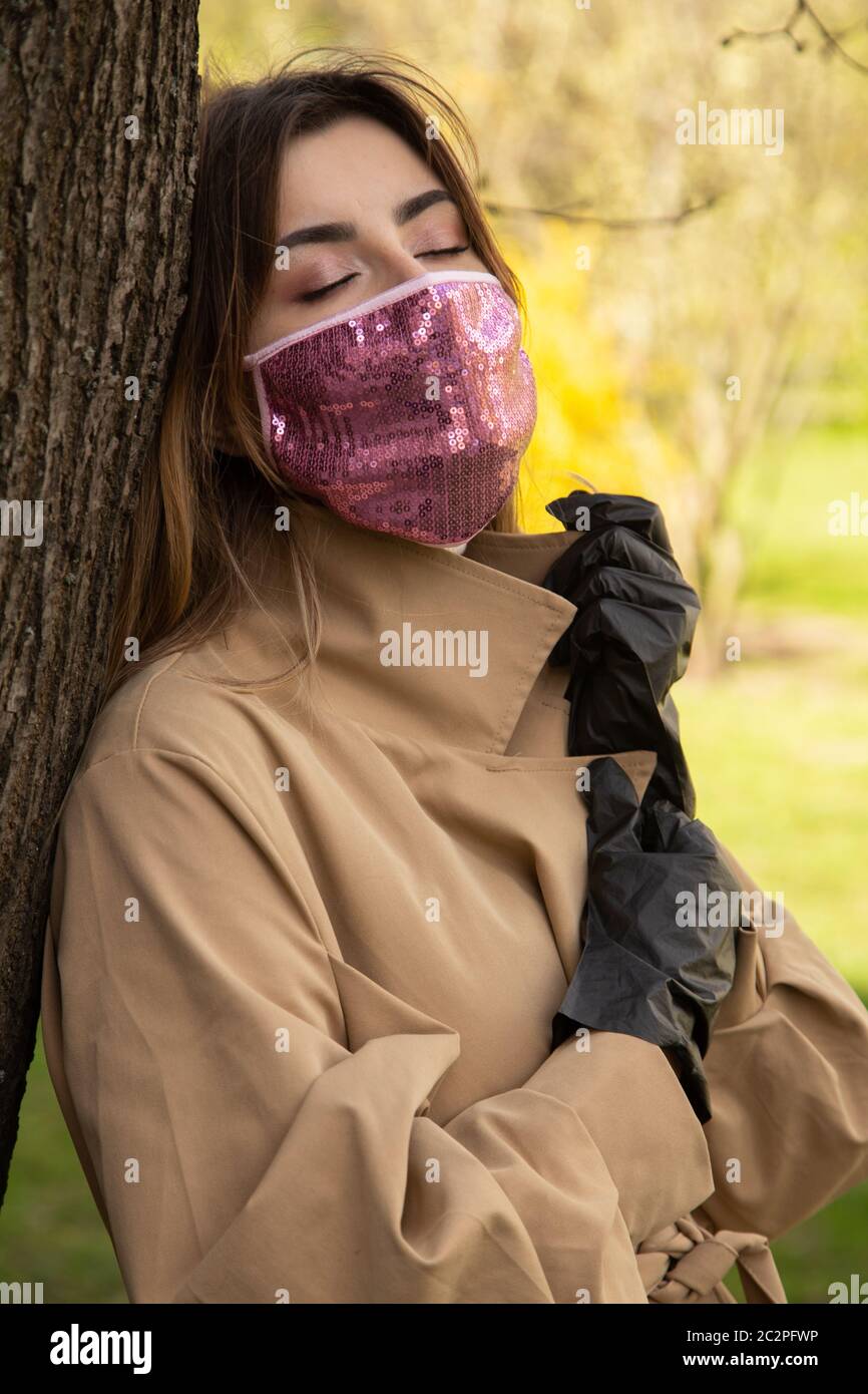 triste giovane donna con maschera medica protettiva con occhi chiusi rilassarsi vicino albero Foto Stock