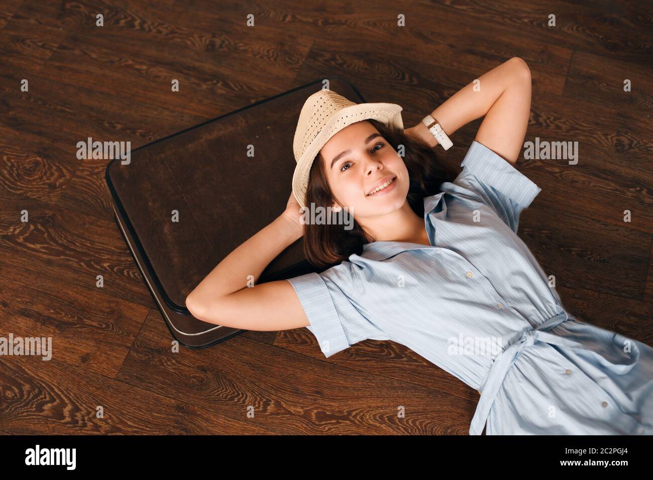 Sorridente ragazza in azzurro vestito e cappello bianco messo la sua testa su valigia e sogni di un viaggio futuro. Ragazza si trova su pavimento di legno. Viaggio in attesa conc Foto Stock