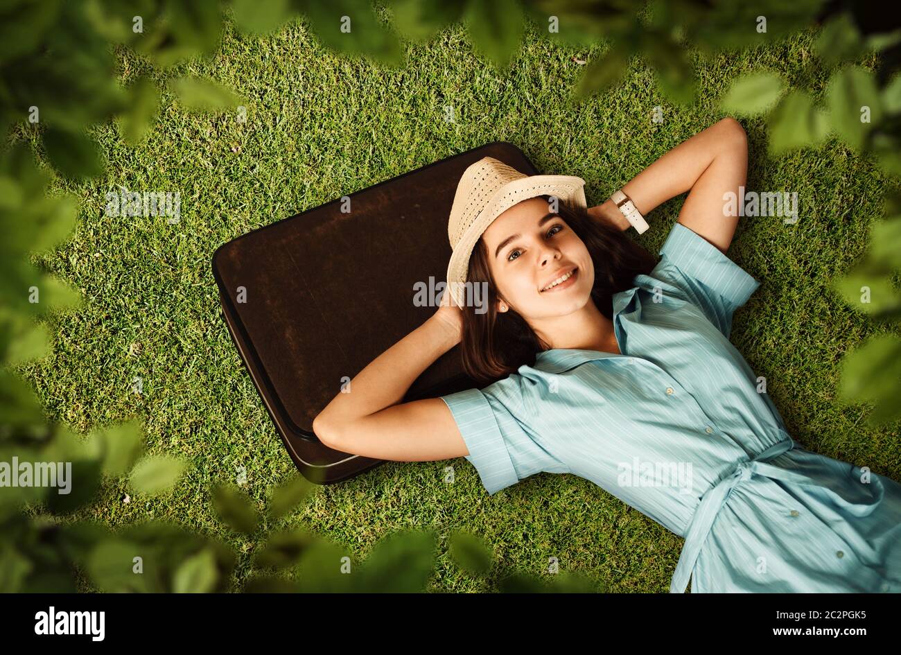 Giovane donna in un abito azzurro e cappello bianco ha messo la testa sulla valigia e sogni di un viaggio futuro. Ragazza giace su erba verde. Viaggio in attesa concep Foto Stock