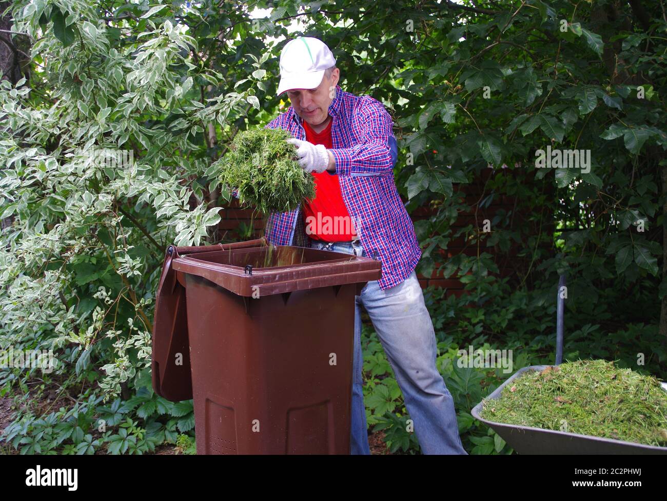 Segregazione dei rifiuti verdi. Un giardiniere gettando fieno verde in un bidone marrone di riciclaggio biologico. Ecologia e l'ambiente nel giardino della casa. Foto Stock