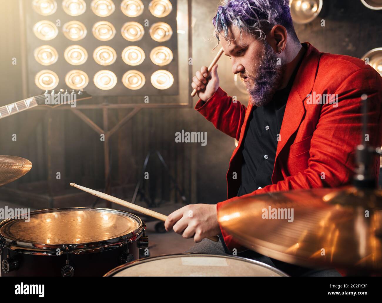 Batterista barbuto in tuta rossa ad esibirsi sul palco con luci, in stile retrò. Esecutore musicale con i capelli colorati, strumento drum Foto Stock