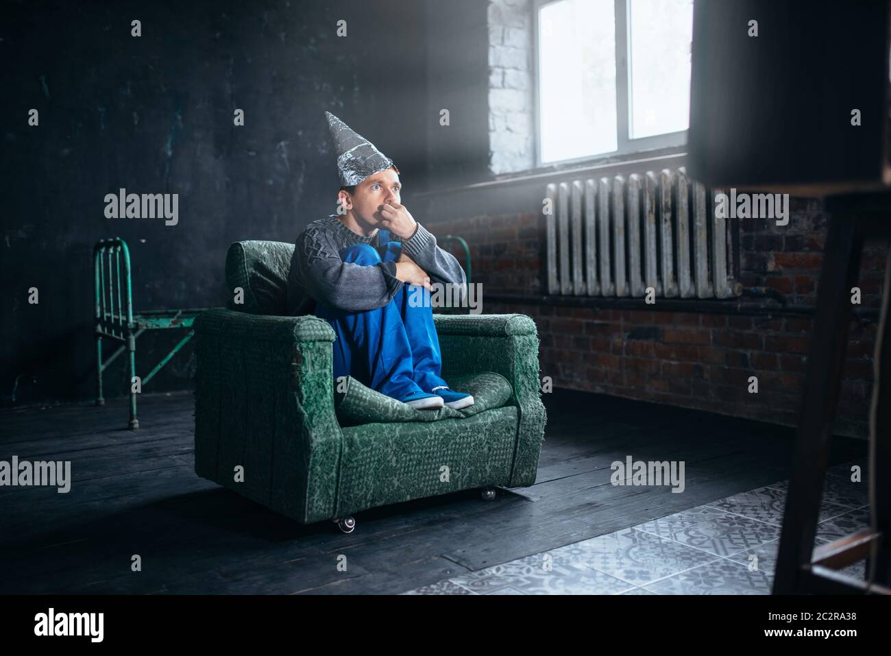 Stupido uomo in tufoil casco guardare TV, paranoia concetto. UFO, teoria della cospirazione, protezione della telepatia, fobia Foto Stock