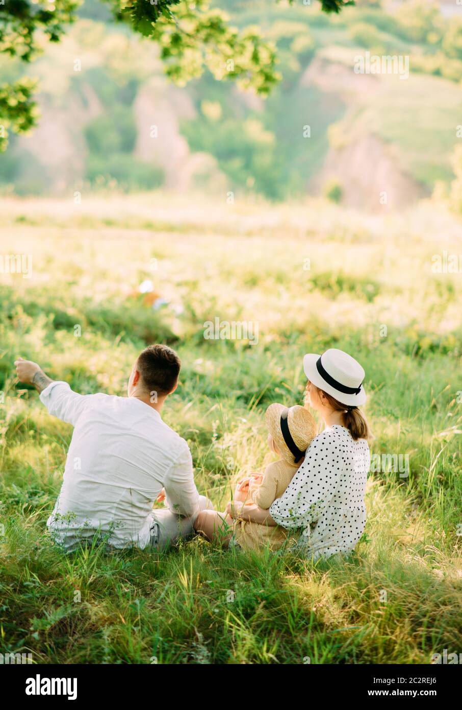I giovani genitori siedono sull'erba con la figlia mentre camminano nel parco. Vista posteriore. Foto Stock