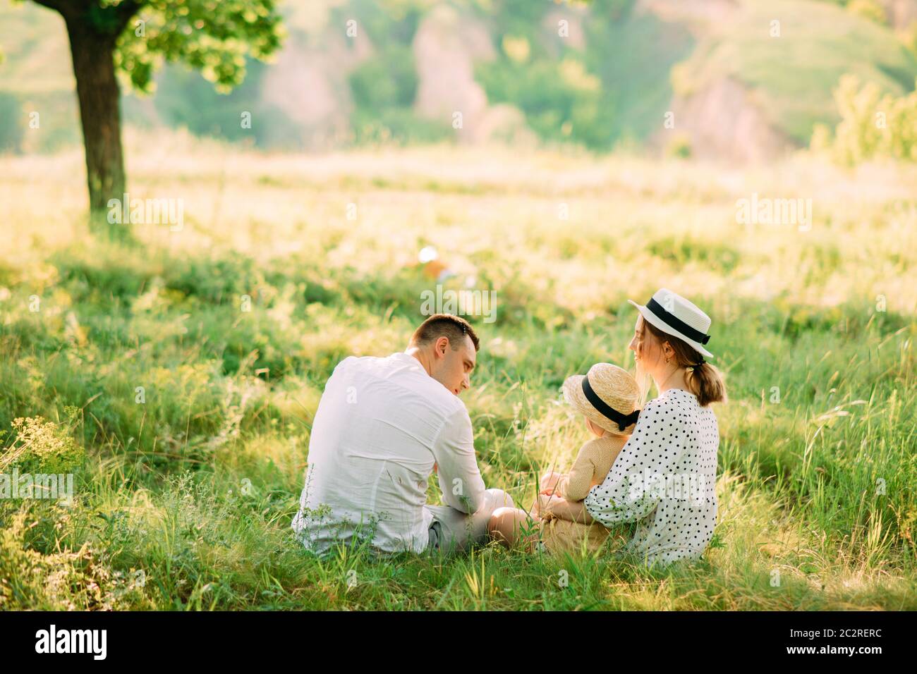 I giovani genitori siedono sull'erba con la figlia mentre camminano nel parco. Vista posteriore. Foto Stock