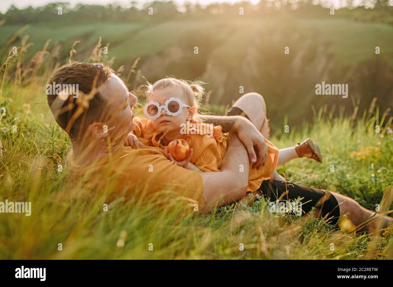 Il giovane padre giace in erba e abbraccia sua figlia mentre cammina nel parco. Foto Stock