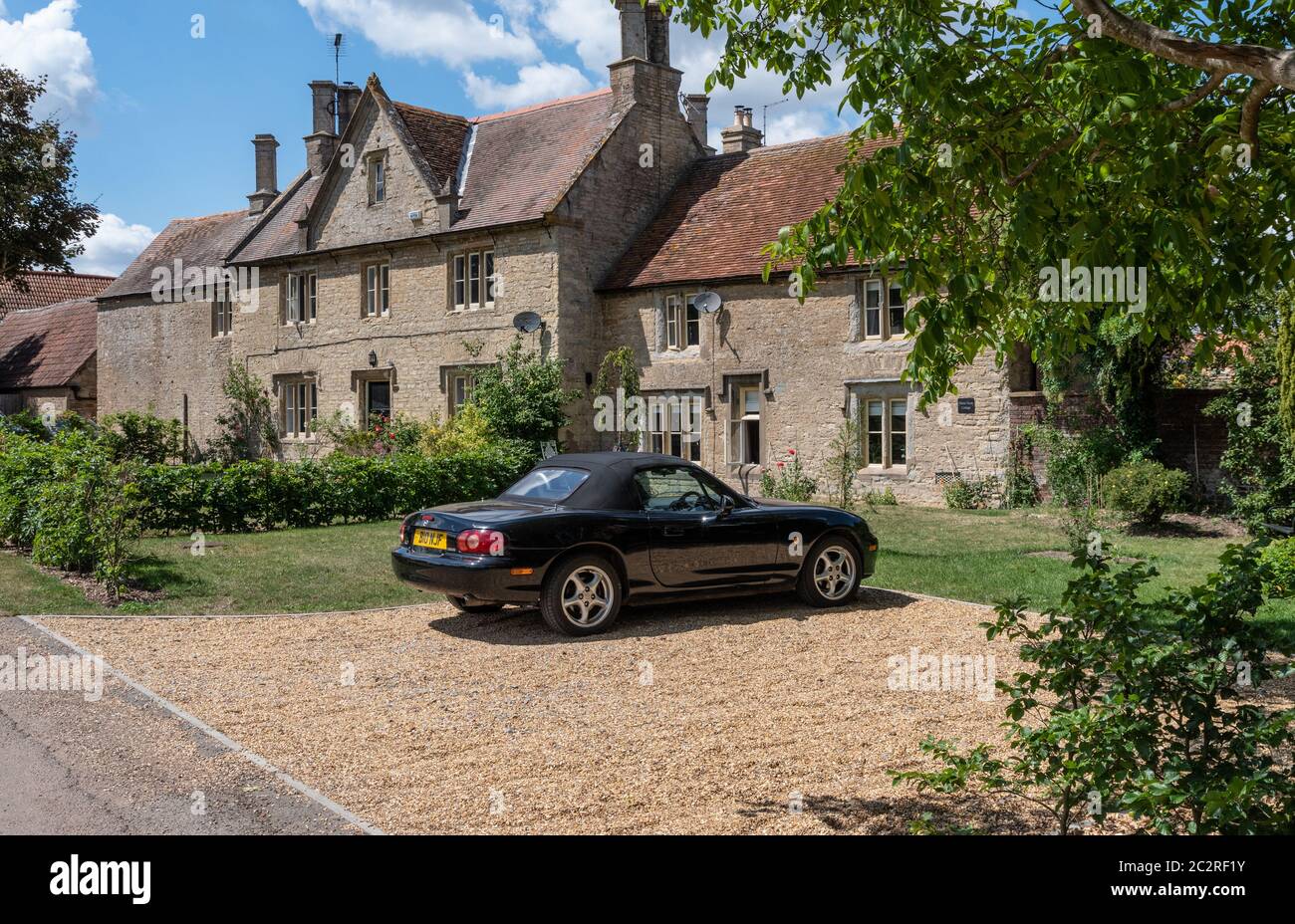 Una scena tradizionale inglese paese, una macchina sportiva parcheggiata di fronte a vecchi cottage in pietra; Castle Ashby, Northamptonshire, Regno Unito Foto Stock