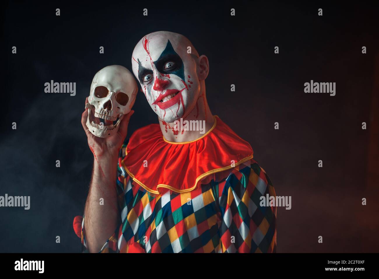 Bloody clown con crazy face detiene il teschio umano, terribile segreto. L'uomo con il trucco in costume di carnevale, mad maniac Foto Stock