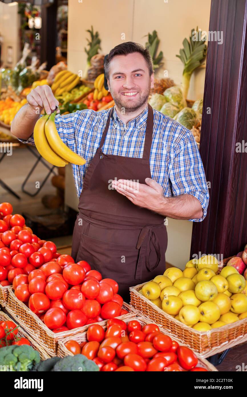 L'uomo sorridente offre banane fresche davanti a scatole con pomodori e mele gialle. Negozio di alimentari sullo sfondo. Foto Stock
