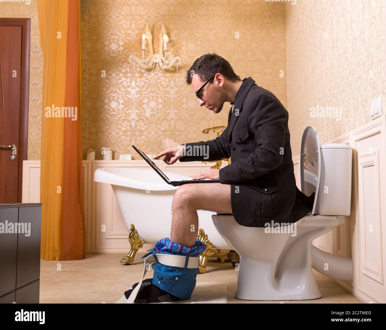 Uomo in bicchieri con laptop seduto sulla tazza di gabinetto. Lussuoso bagno interno in stile vintage sullo sfondo Foto Stock