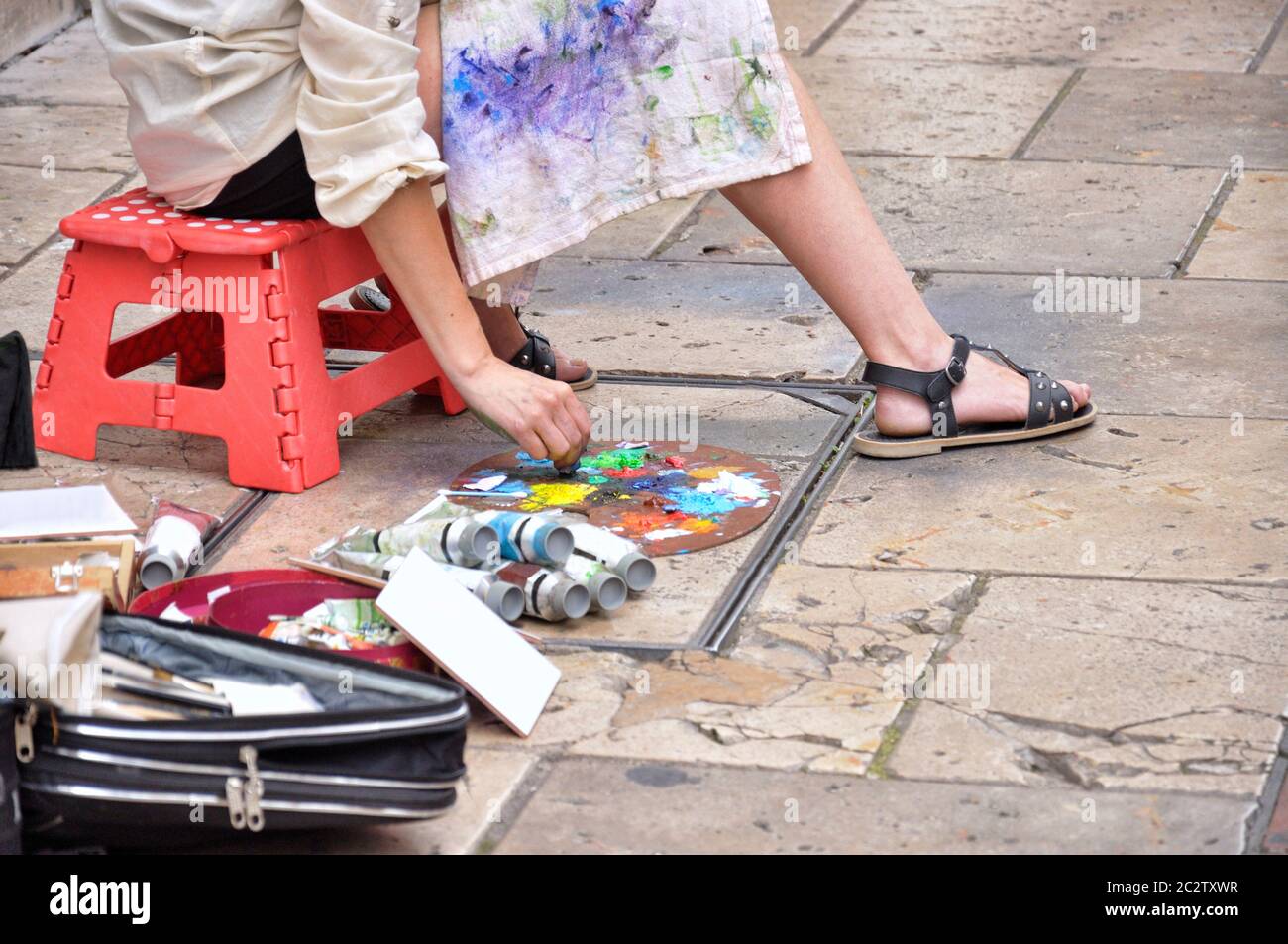 Donna artista pittura sulla strada, è seduta su una panca rossa, attrezzi dipingono accanto ai piedi. Foto Stock