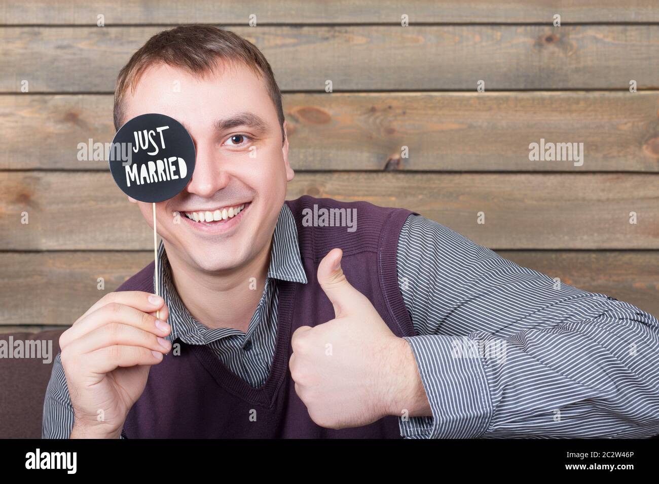 Giovane uomo detiene divertente icona su un bastone con appena sposati Iscrizione e mostrare pollice in alto segno, sullo sfondo di legno. Fun photo props e accessori per p Foto Stock