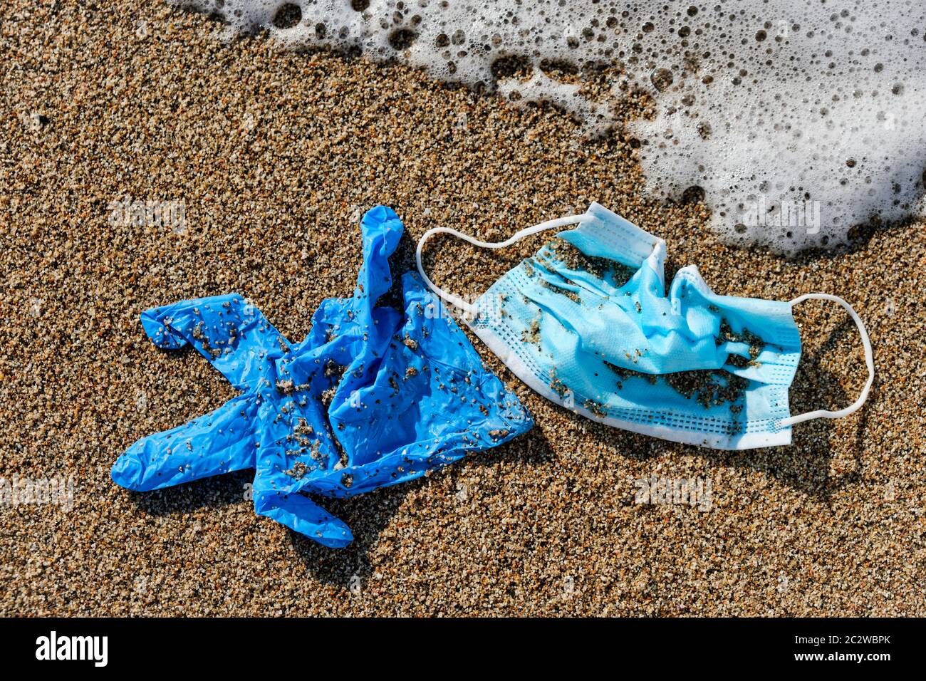 primo piano di una maschera chirurgica blu usata e un guanto di lattice blu gettato sulla sabbia bagnata della riva di una spiaggia Foto Stock