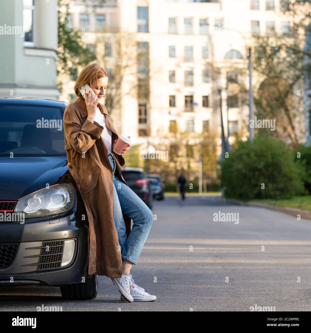 Giovane donna caucasica in piedi vicino alla sua auto e parlando al telefono, tenendo una tazza di caffè, indossando trench beige Foto Stock