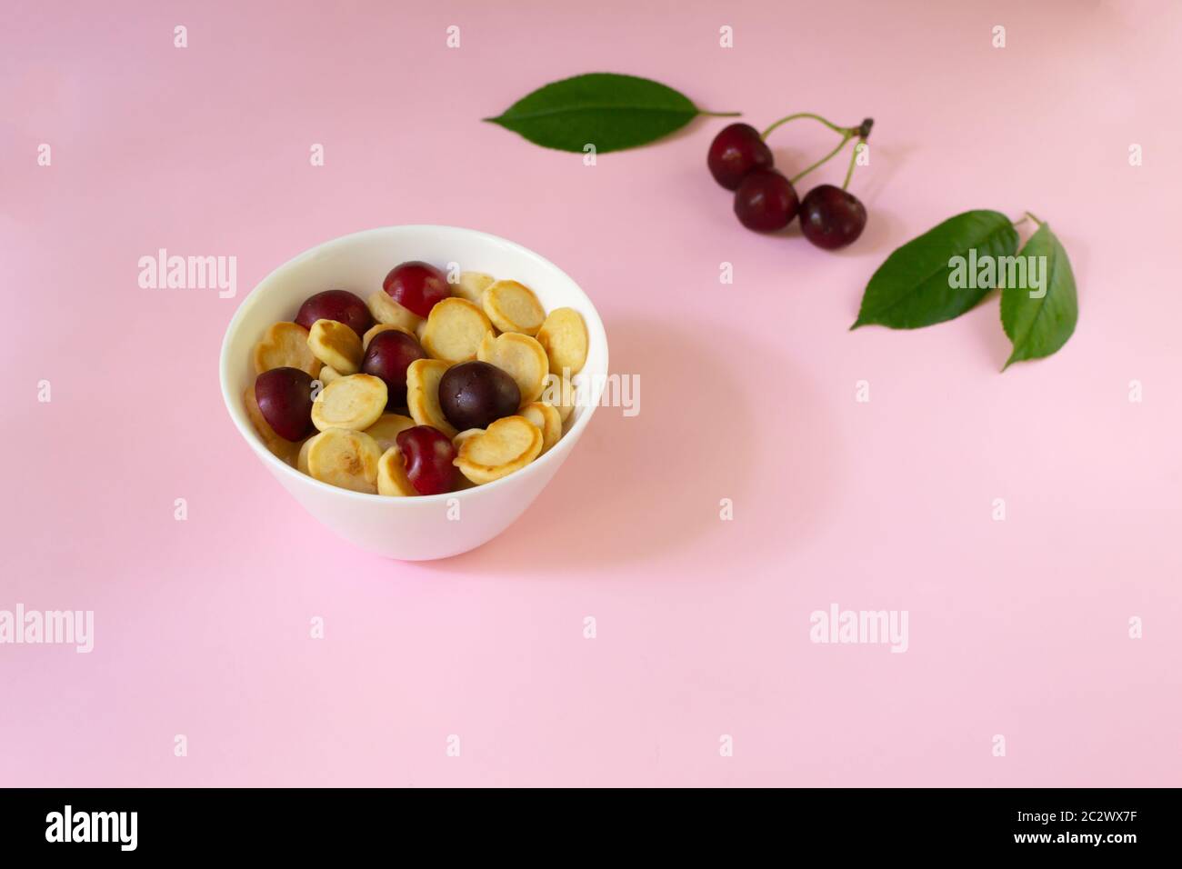 Una ciotola bianca di cereali con un piccolo pancake con ciliegie, bacche e foglie verdi sullo sfondo su sfondo rosa. Spazio di copia. Cucina alla moda con Foto Stock