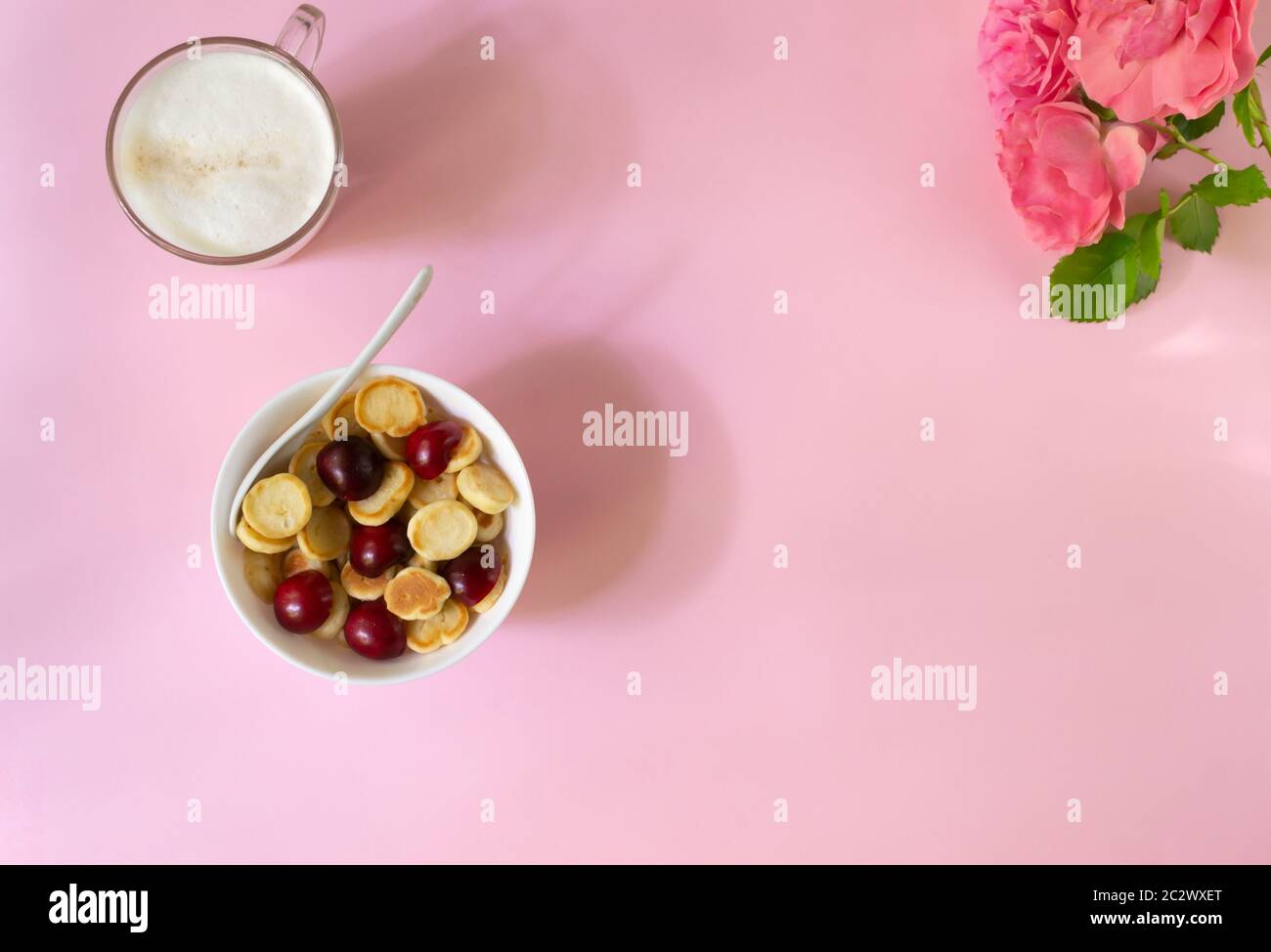 Una ciotola bianca di cereali con una piccola frittella con ciliegie, una tazza di cappuccino e fiori su sfondo rosa. Spazio di copia. Vista dall'alto. Cucina alla moda con Foto Stock