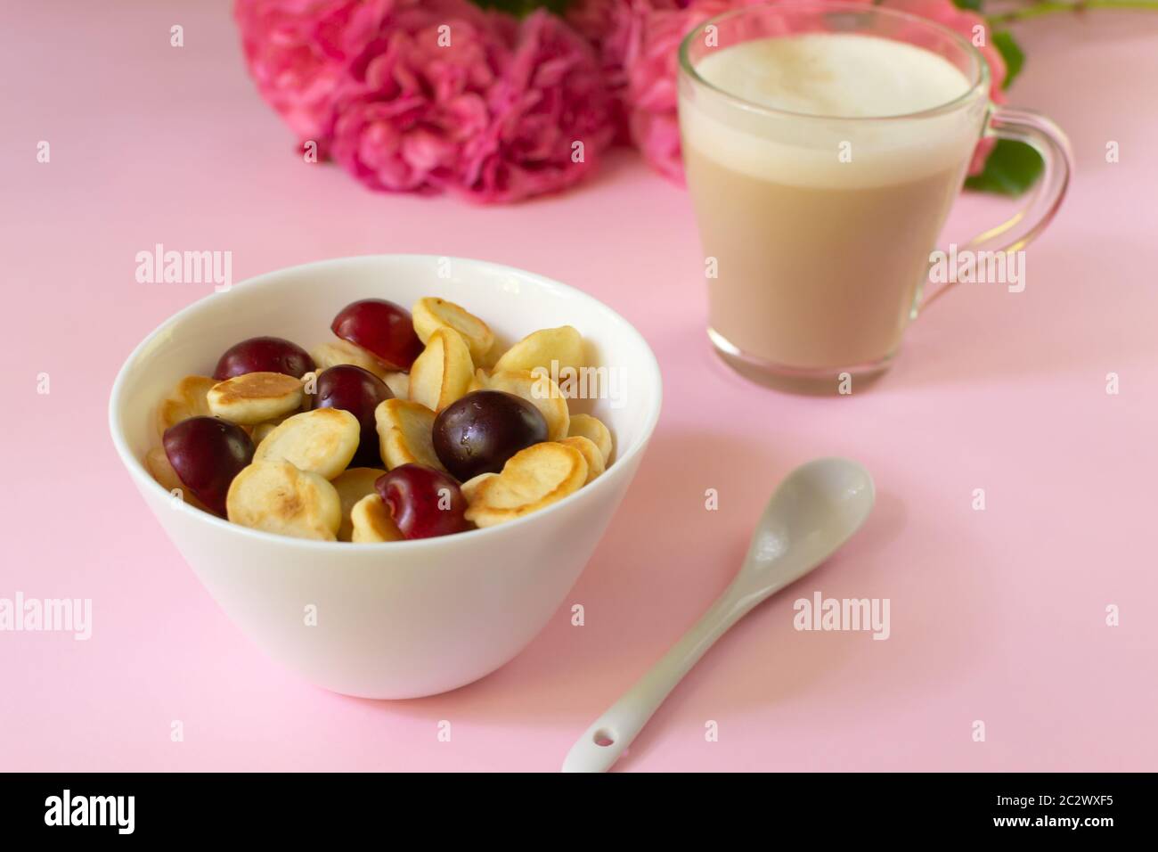 Una ciotola bianca di cereali con una piccola frittella con ciliegie, una tazza di cappuccino e fiori su sfondo rosa. Primo piano. Cucina alla moda. Foto Stock