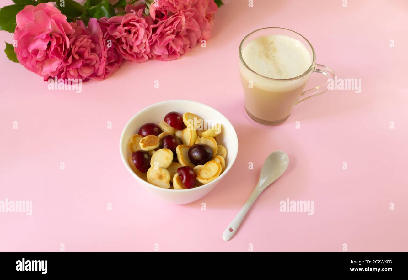 Una ciotola bianca con cereali, una piccola frittella con ciliegie e una tazza di cappuccino, un bouquet di rose su sfondo colorato. Cucina alla moda. Foto Stock