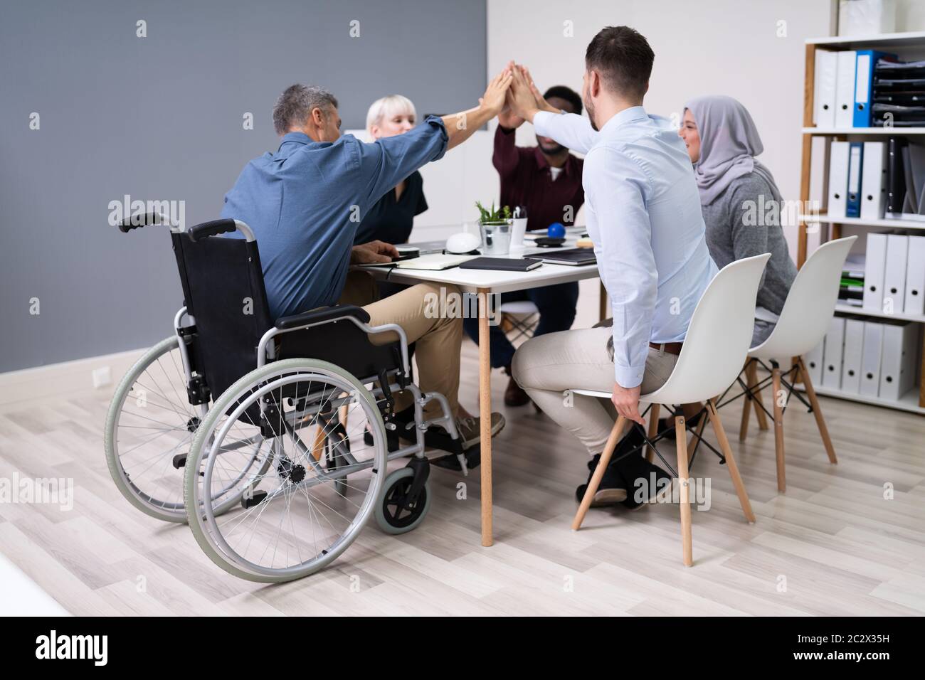 Un Uomo D'Affari Disabile Che Dà Cinque Anni Di Altezza Ai Suoi Partner Sorridenti In Ufficio Foto Stock