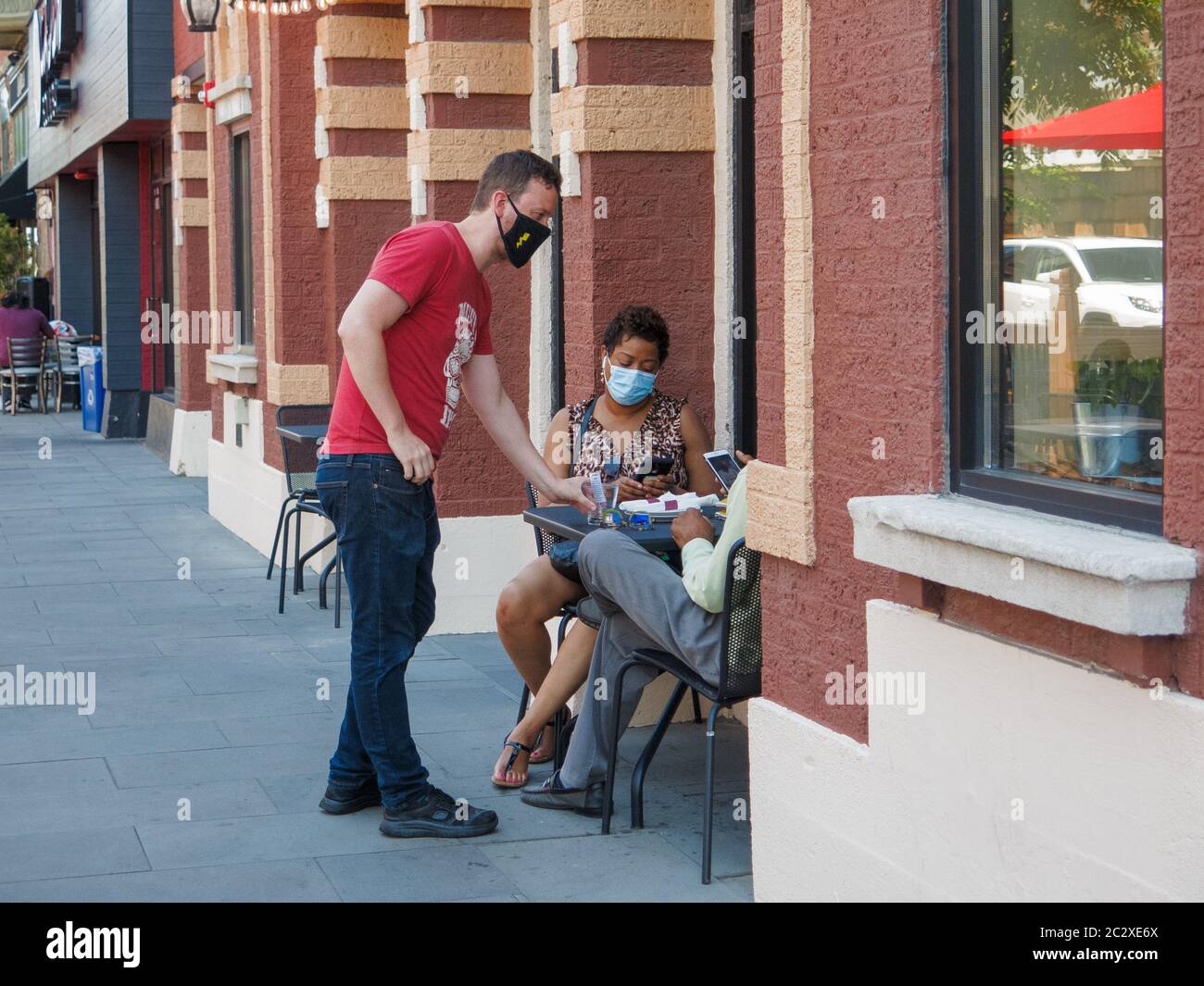 Oak Park, Illinois, Stati Uniti. 17 giugno 2020. Un cameriere serve i clienti in una caffetteria sul marciapiede. I ristoranti dell'area di Chicago sono ora aperti per sedersi all'aperto con un adeguato allontanamento sociale. I server devono indossare maschere facciali. Il tasso di infezione COVID-19 dell'Illinois sta diminuendo più rapidamente di altri stati a causa del soggiorno vigoroso a casa e delle linee guida di distanziamento sociale. Foto Stock