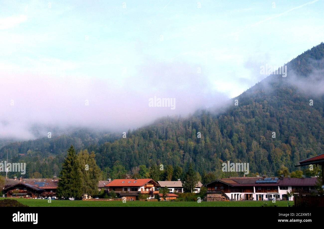 paesaggio con fattorie in primo piano e montagna boscosa dietro con nebbia e cielo blu Foto Stock