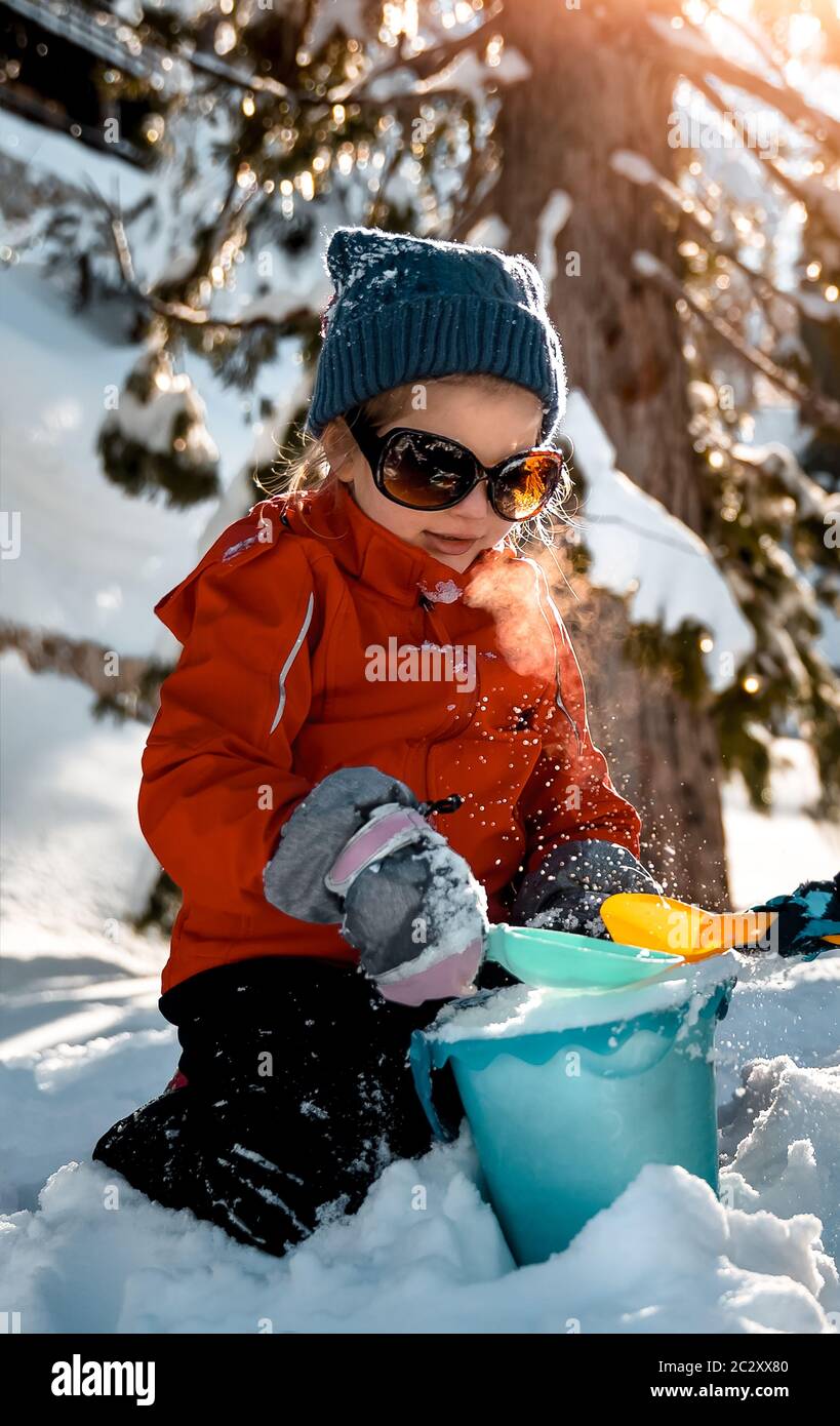 Bambina che si gode vacanze invernali, grazioso bambino vestito con abiti caldi e occhiali da sole divertirsi all'aperto sul cortile e giocare con la neve Foto Stock