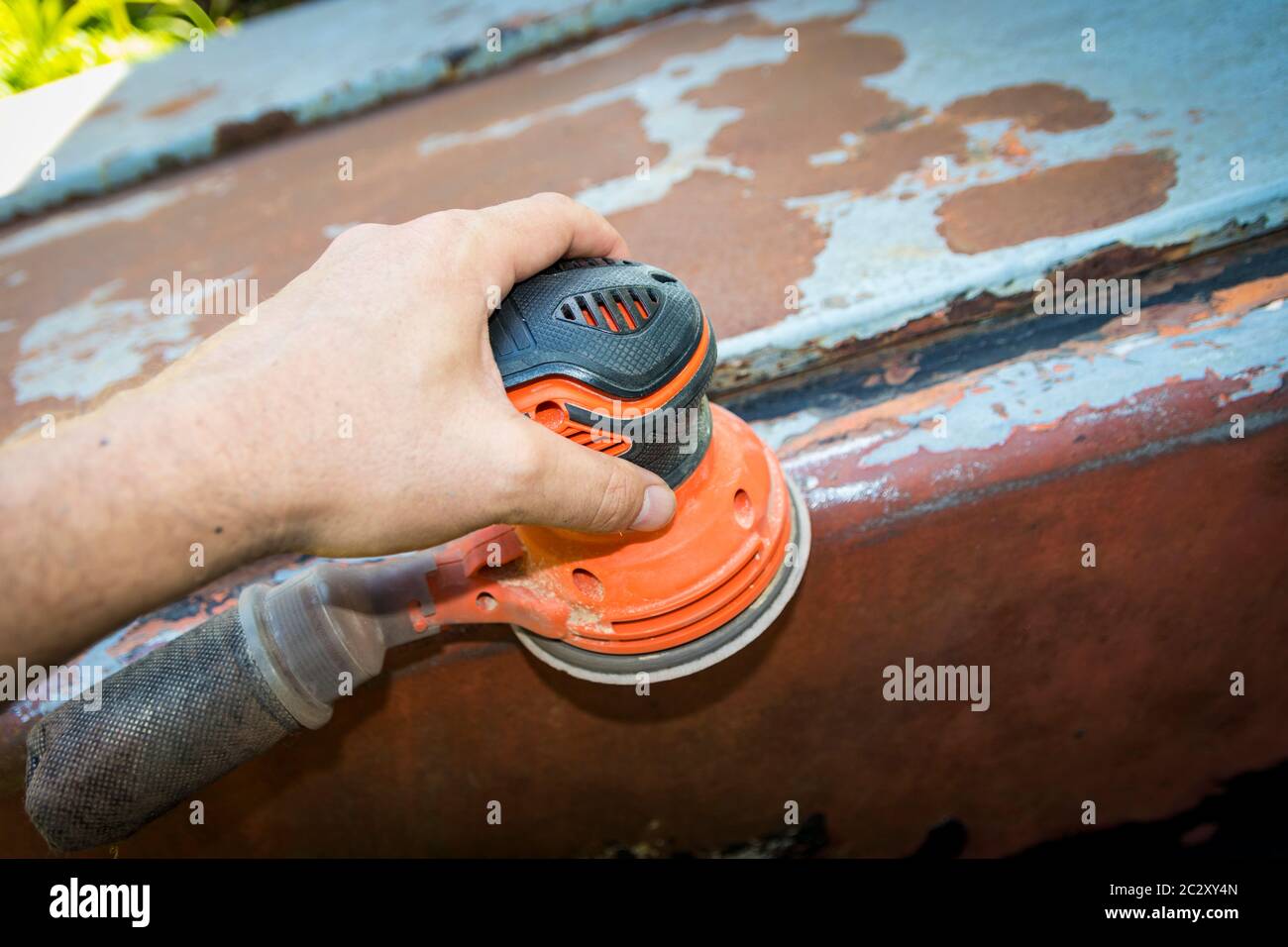 La mano dell'uomo aziona una levigatrice a disco/orbitale, per rimuovere vernice e ruggine dalle porte metalliche Foto Stock