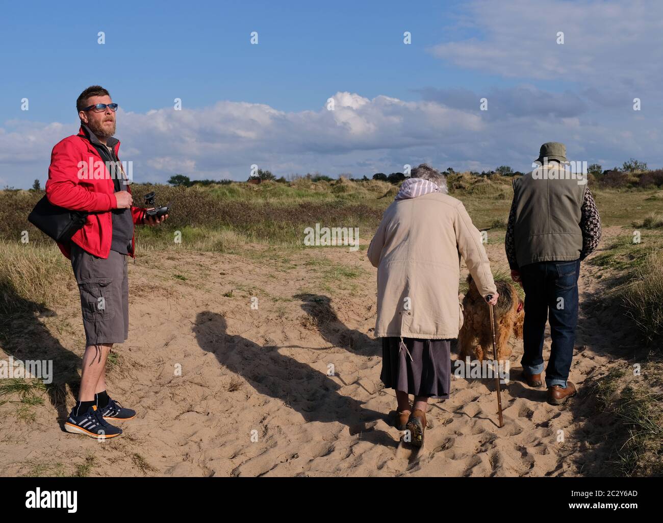 la vecchia coppia con il cane cammina davanti al giovane uomo in giacca rossa guardando verso il mare sul sentiero lato spiaggia in sole luminoso con cielo blu e lunghe ombre Foto Stock