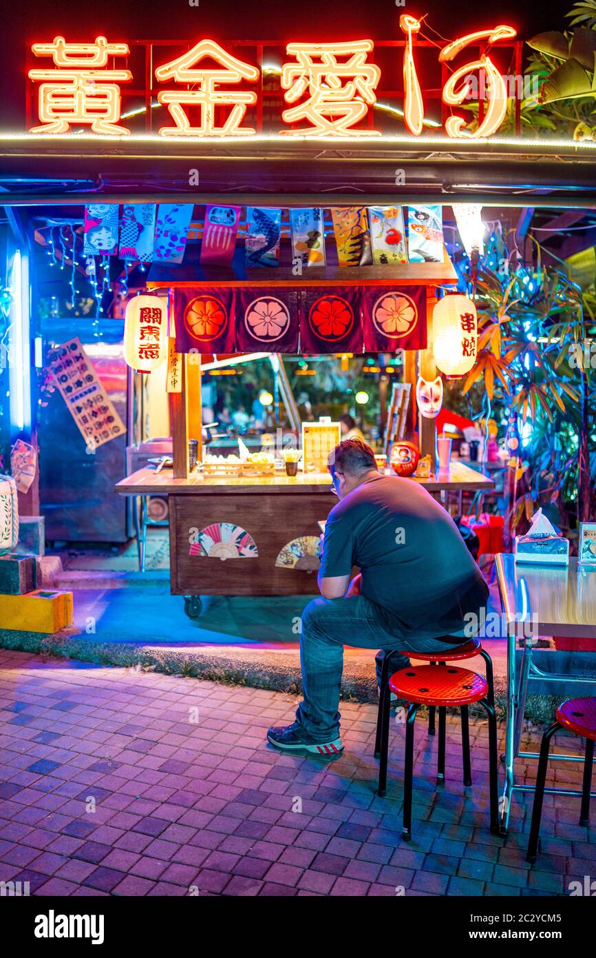 Un uomo che usa il suo smartphone davanti a un colorato, luce al neon coperto piccolo venditore di strada di notte, a Kaohsiung, Taiwan Foto Stock