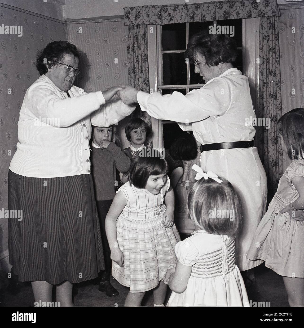 Anni '60, storico, all'interno di una stanza di un bambino festa di compleanno, eccitato giovani di divertirsi giocando un gioco che passa sotto le braccia unite di due Signore, Inghilterra, Regno Unito. Foto Stock