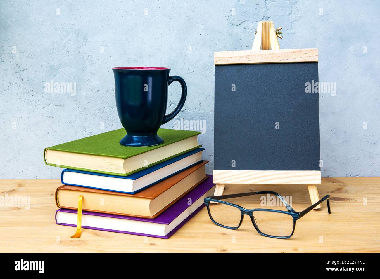 piccola lavagna su tavolo di legno con libri, coppa e occhiali con sfondo grigio Foto Stock
