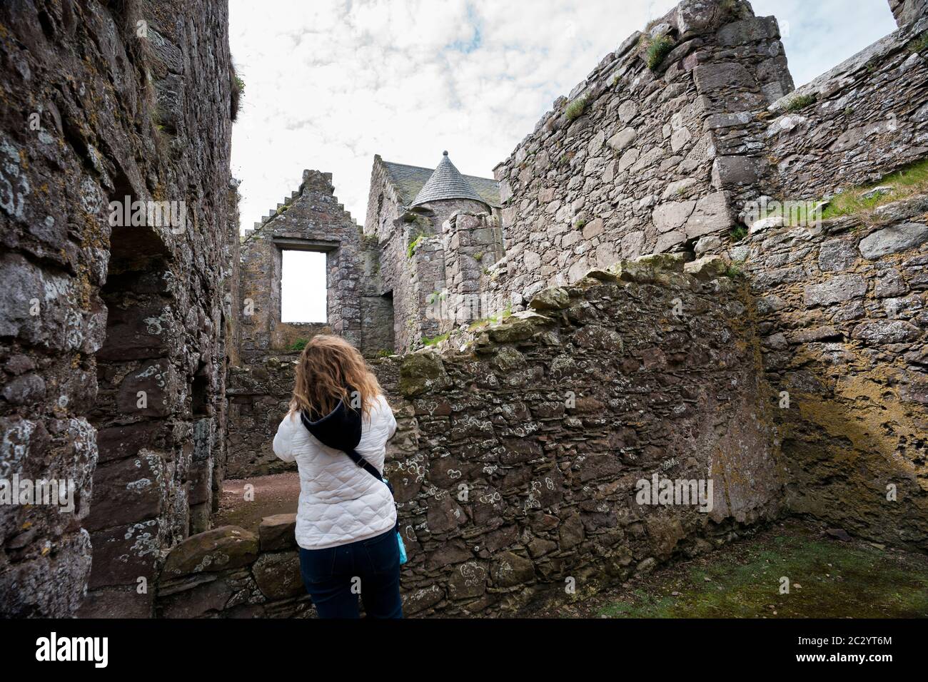 Un turista biondo esplora le mura che si sbriciolavano sotto un cielo nuvoloso nel castello in pietra rovinato di Dunnottar, Stonehaven, Scozia, Regno Unito Foto Stock