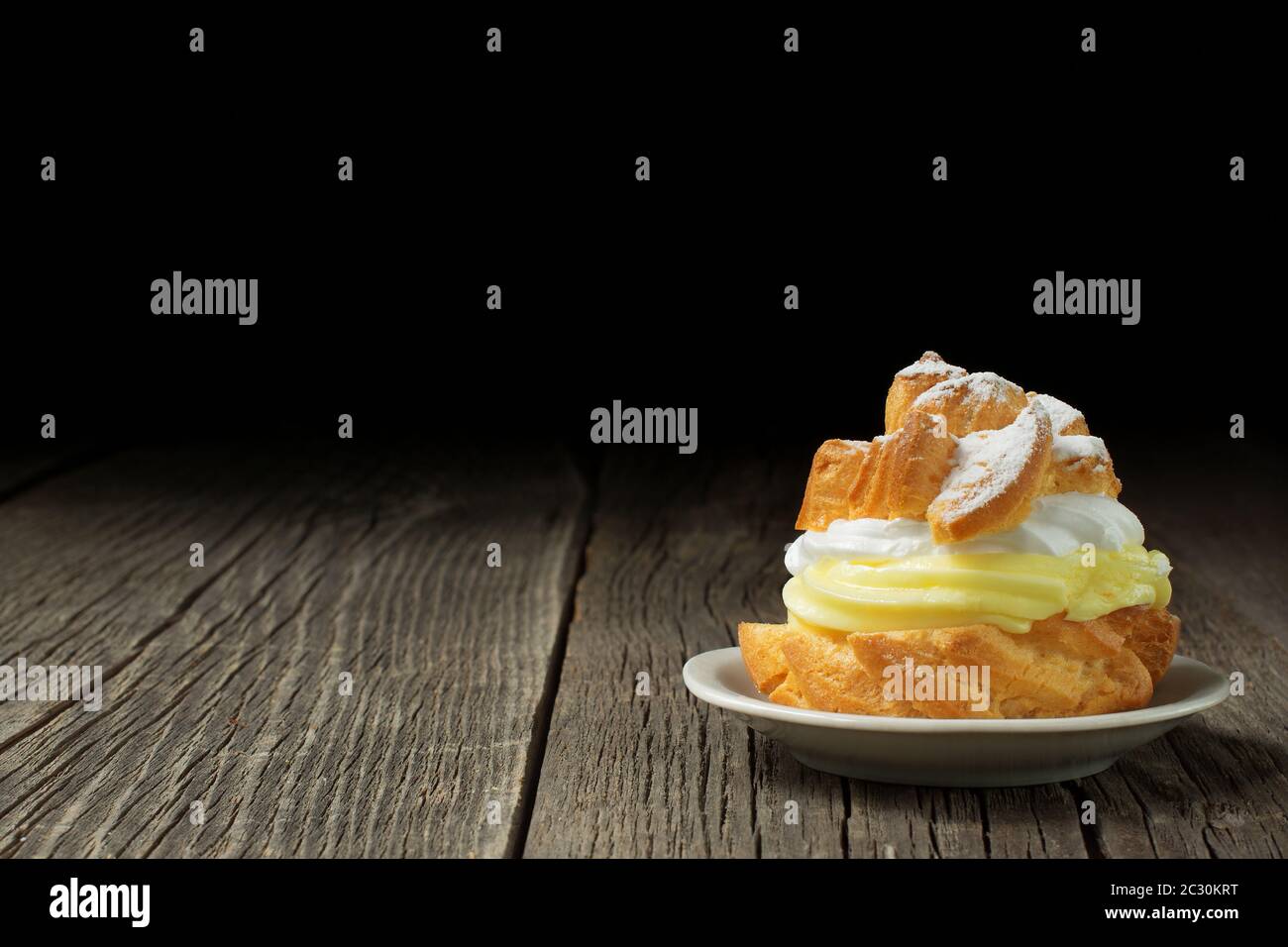 Un profiterolo, decorato con uno spolverato di zucchero in polvere e riempito con abbondante crema di crema, è adagiato su un piccolo piatto di ceramica. Foto Stock