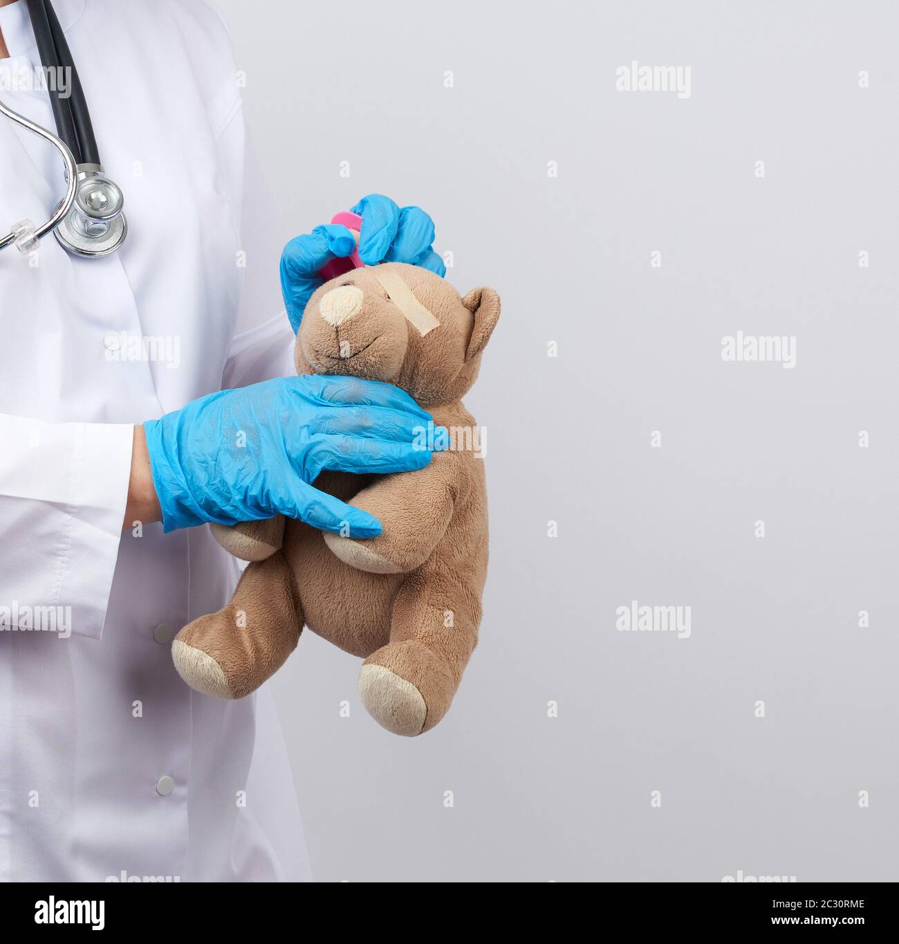il medico in un cappotto bianco e guanti di lattice blu tiene un orsacchiotto marrone senza occhio, il medico avvolge la sua testa con cerotto adesivo Foto Stock