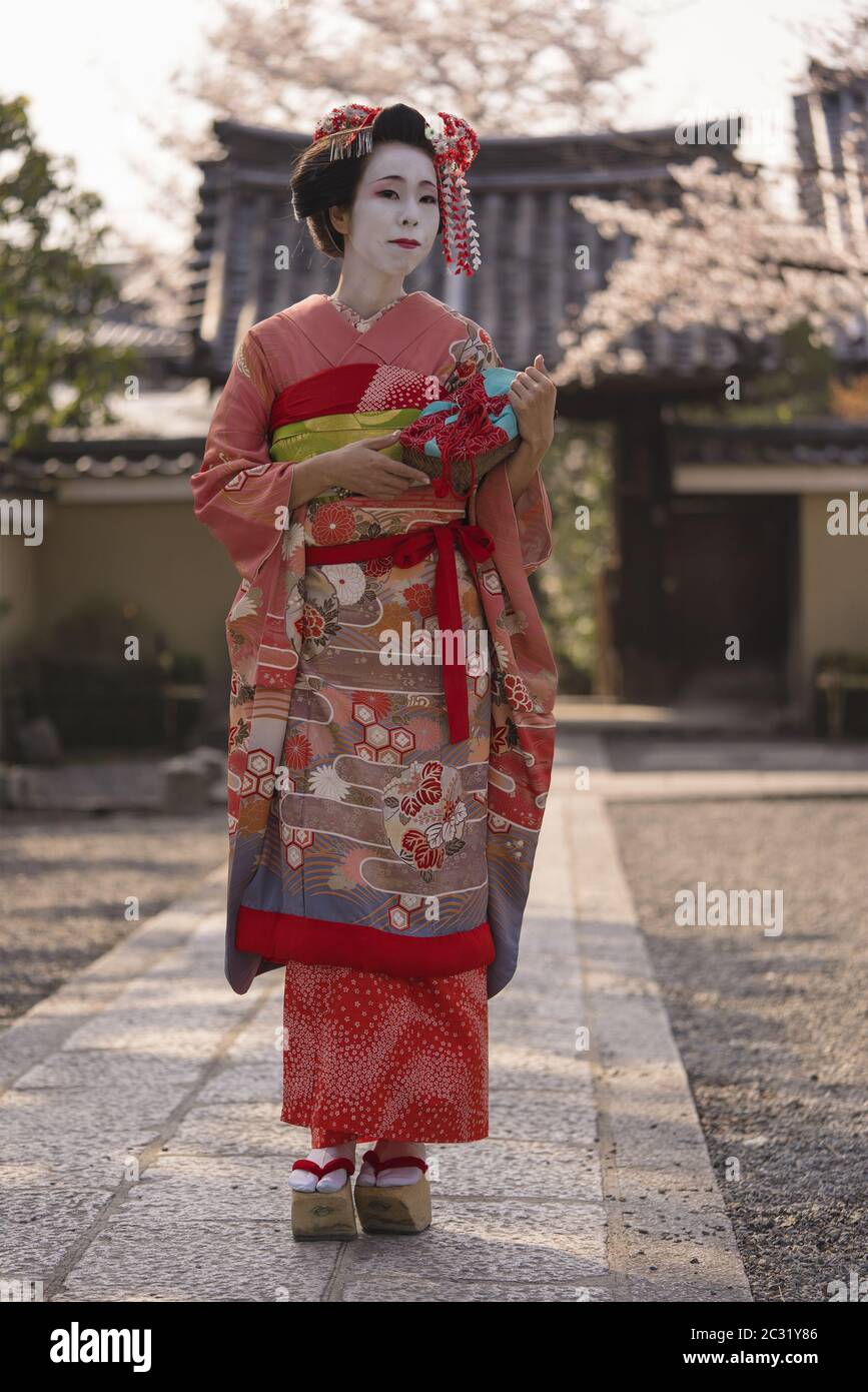 Maiko in un kimono a piedi su un sentiero in pietra di fronte al cancello di un tempio tradizionale giapponese Foto Stock