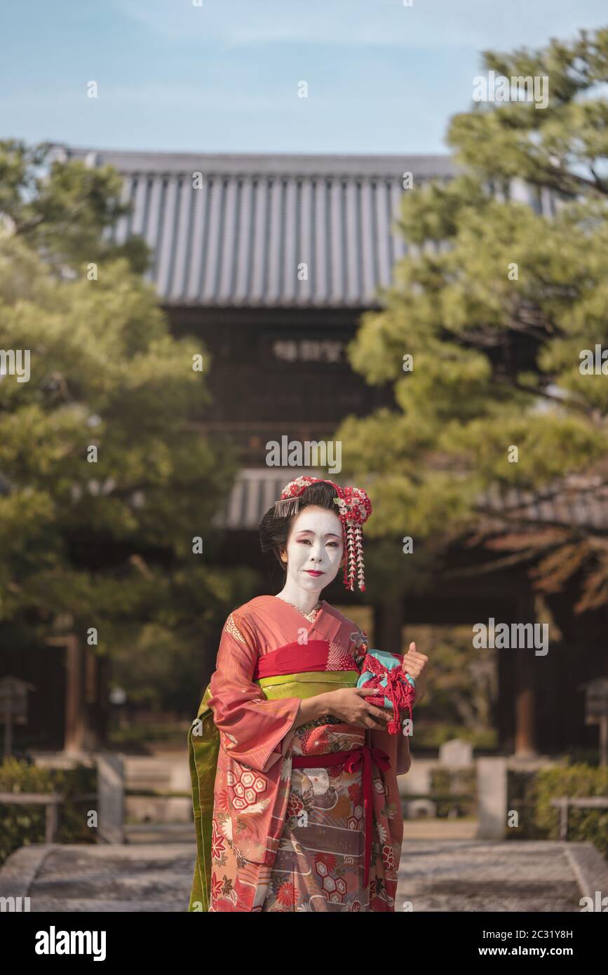Maiko in un kimono che si posa a Kyoto su un ponte di pietra di fronte alla porta di un tempio giapponese Foto Stock