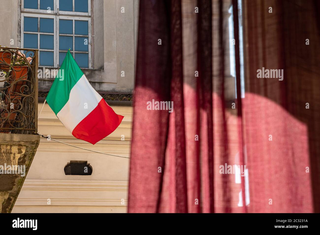 Bandiera italiana che si affaccia sul vento su un balcone in una giornata di sole. Guardando fuori da una finestra durante la chiusura a causa di un focolaio di coronavirus. Foto Stock