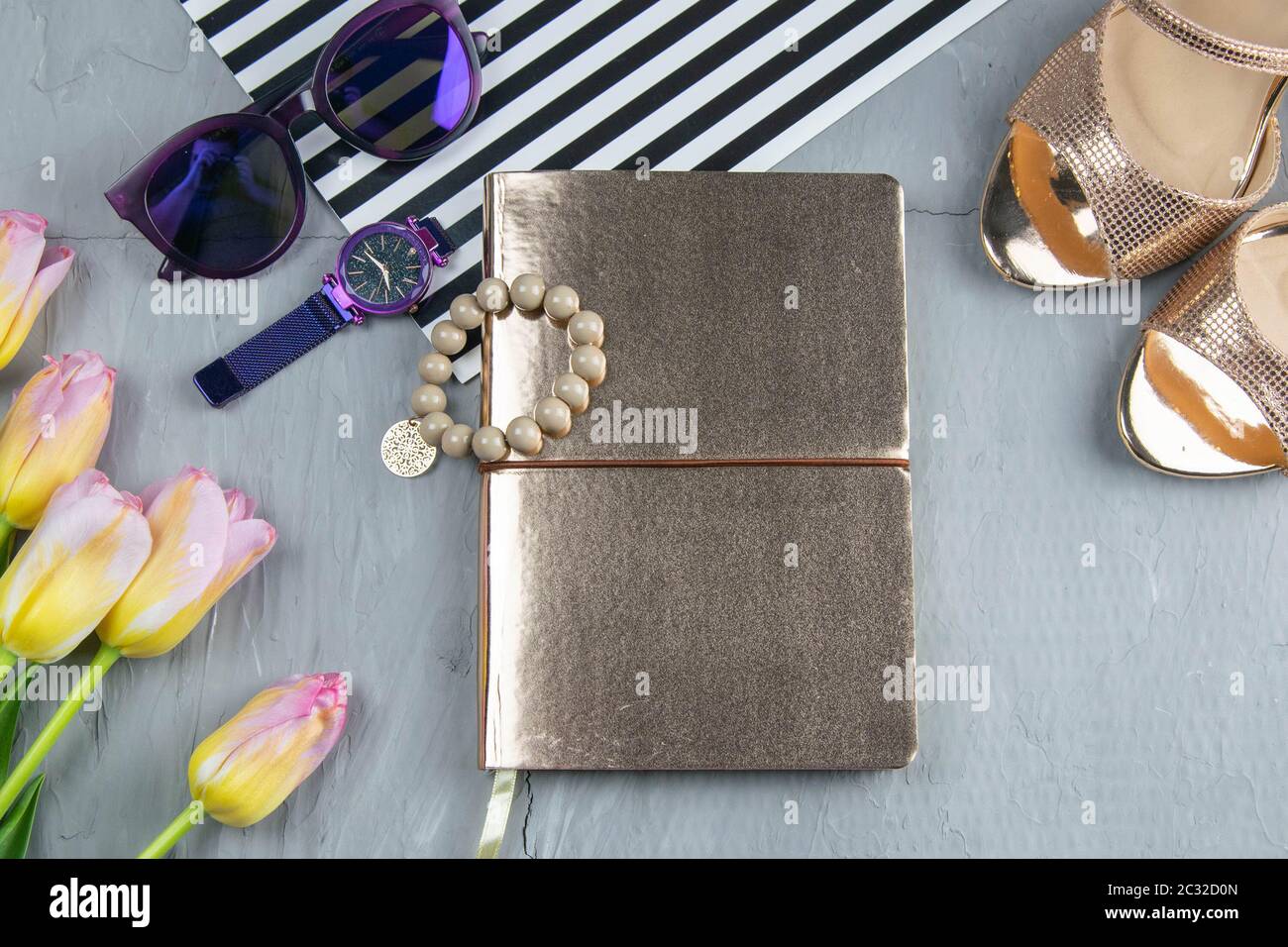 Un taccuino d'oro con scarpe da donna, tulipani, occhiali e un orologio su sfondo di cemento. Posa flet. Foto Stock