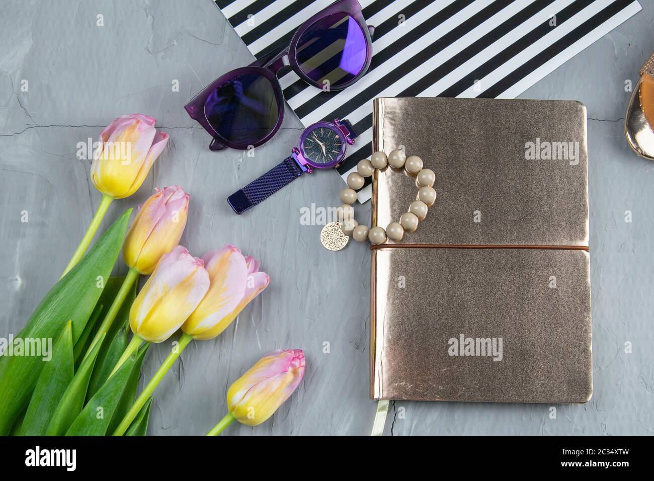 Un taccuino d'oro con scarpe da donna, tulipani, occhiali e un orologio su sfondo di cemento. Posa flet. Foto Stock