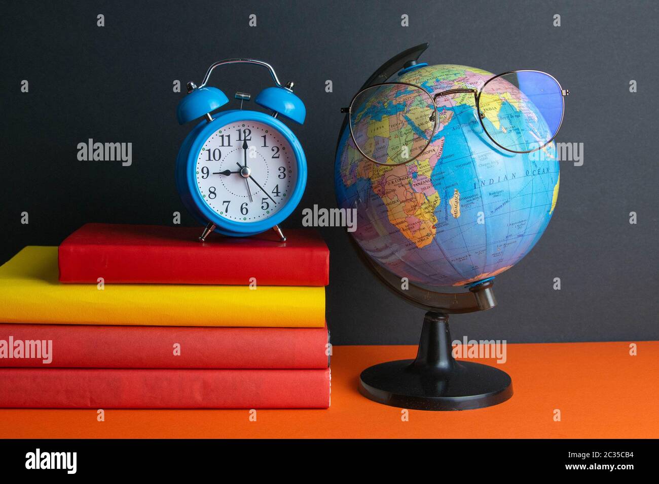 Una pila di libri, orologio, globo mondiale e occhiali da donna in primo piano, si trovano su uno sfondo arancione. Foto Stock