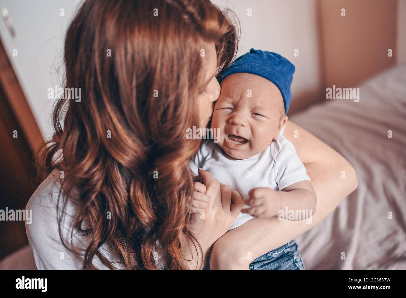 Primo piano ritratto di felice mamma giovane che tiene il suo bambino piangente in jeans blu e t-shirt bianca e cappuccio. Giovane famiglia felice, madre che gioca con cute Foto Stock