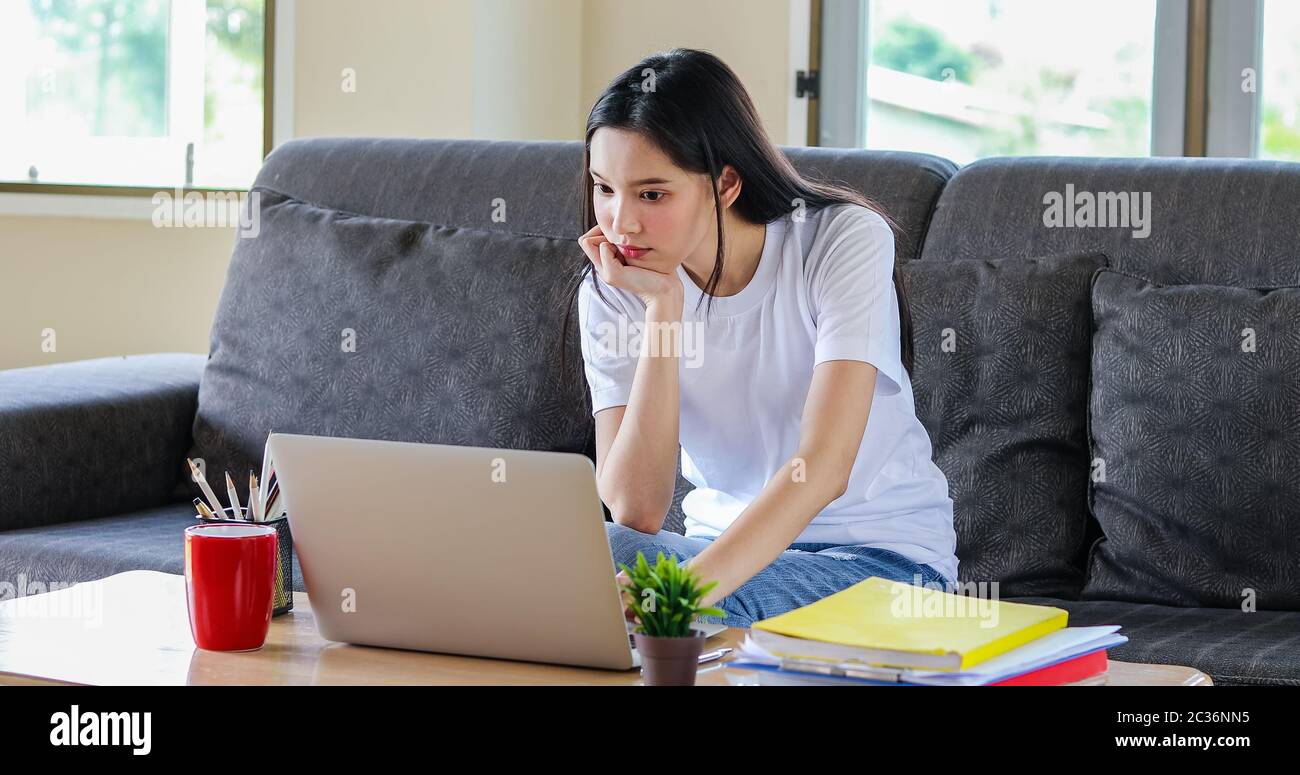 Donna asiatica felice allegra cute bello sta usando un computer del notebook e seduta sul divano per lavorare da casa. Durante il Th è messa in quarantena a casa Foto Stock