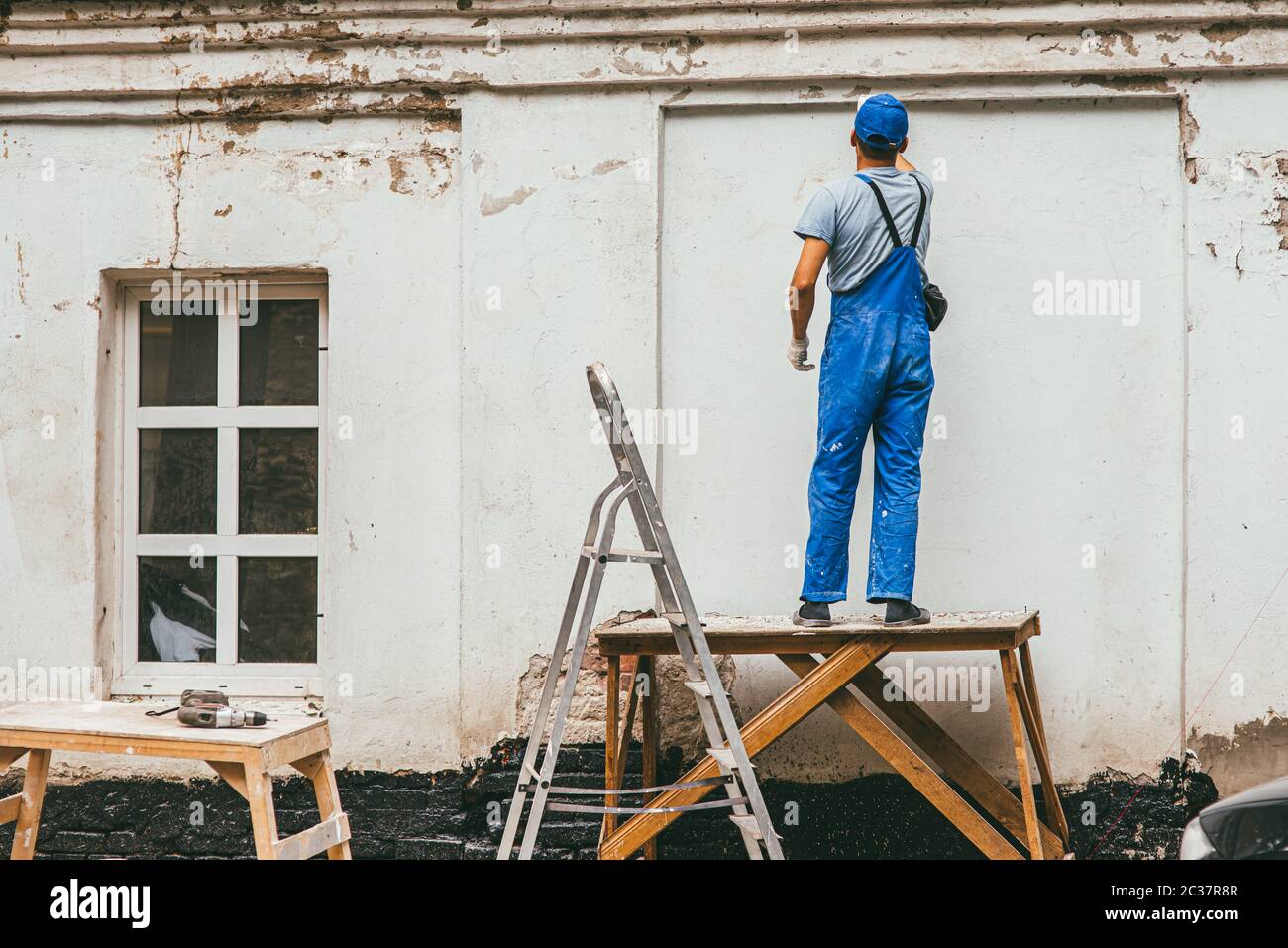 lavoratore irriconoscibile in blu pantaloni costruzione si trova su un stand di legno e lavora con una spatola con intonaco sulla parete. Ren Foto Stock