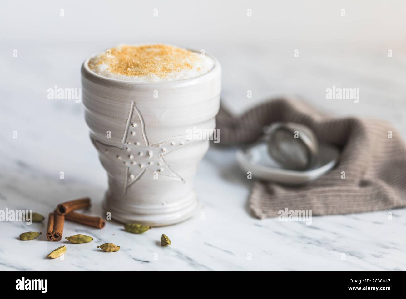 Una tazza di latte di chai con zucchero di canna sulla parte superiore. Su un tavolo di marmo con sfondo bianco. Ci sono alcuni cialde di cardamomo e bastoncini di cannella accanto alla whit Foto Stock